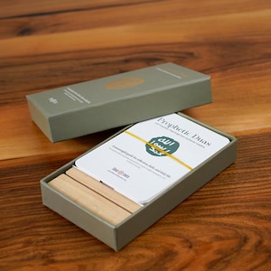 K&ouml;nnte beinhalten: Ein Satz Prophetic Duas-Karten in einer grauen Box mit passendem Deckel. Die Karten sind wei&szlig; mit Text und einem gr&uuml;nen Emblem. Zwei Holzbl&ouml;cke befinden sich in der Box. Die Box steht auf einer Holzoberfl&auml;che.