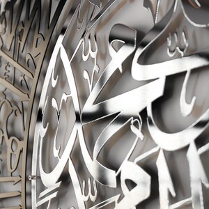 May include: A silver metal wall decor with intricate Arabic calligraphy. The design is cut out of the metal, creating a three-dimensional effect.