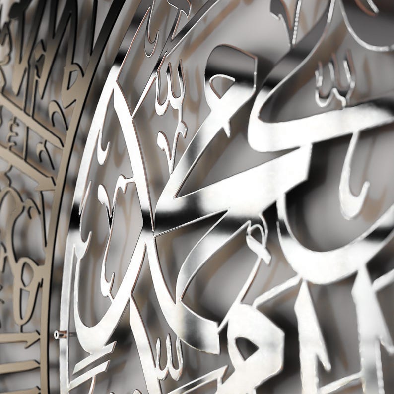 May include: A silver metal wall decor with intricate Arabic calligraphy. The design is cut out of the metal, creating a three-dimensional effect.