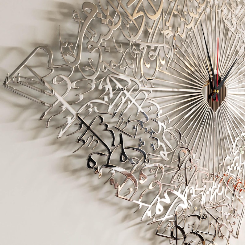 May include: A silver-colored wall clock with a unique design featuring Arabic calligraphy. The clock has a radial pattern with thin lines emanating from the center, where the clock hands are located. The hands are black with a red second hand.