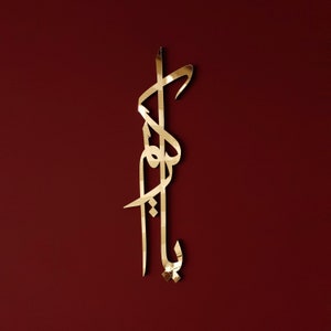 May include: Gold-coloured metal wall art featuring a decorative Arabic calligraphy design. The design is intricate and features flowing lines and curves.