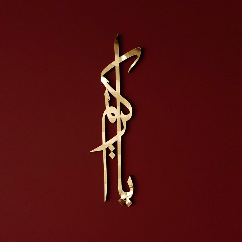 May include: Gold-coloured metal wall art featuring a decorative Arabic calligraphy design. The design is intricate and features flowing lines and curves.