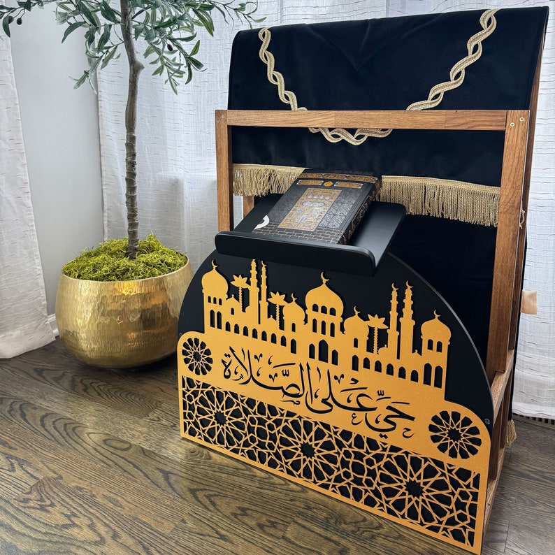 May include: A wooden prayer stand with a black velvet cushion and a gold-colored metal design featuring a mosque and Arabic calligraphy. The stand has a shelf for a book, and the cushion is fringed with gold tassels.
