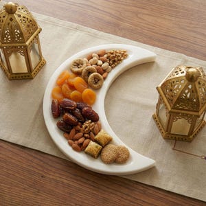 May include: A white crescent-shaped serving dish filled with dried fruits and nuts, including dates, apricots, figs, almonds, and walnuts. Two ornate gold lanterns flank the dish on a neutral-colored tablecloth, creating a festive display.