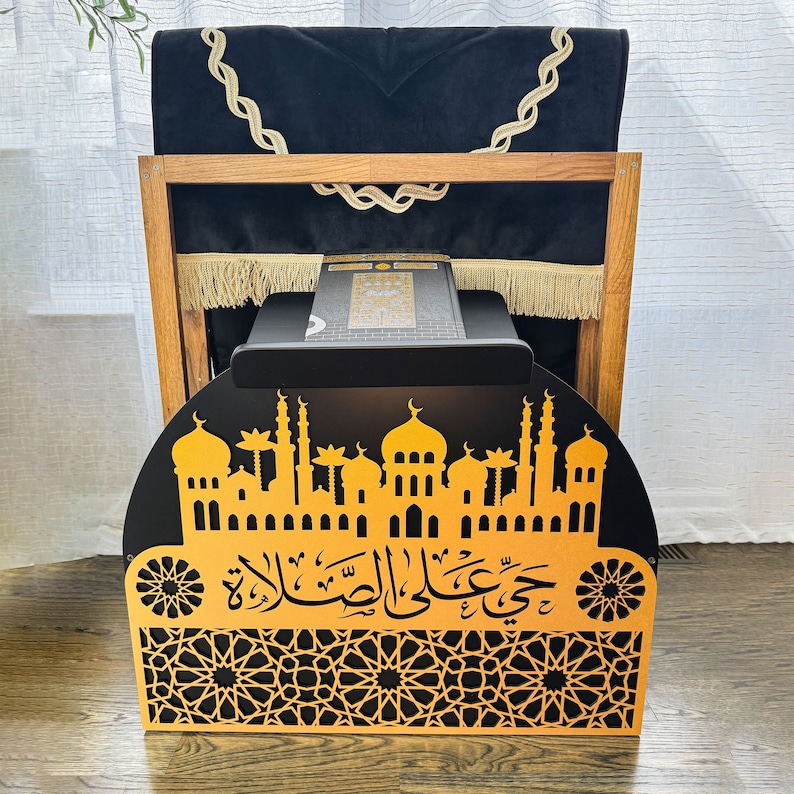 May include: A wooden prayer stand with a black prayer rug and a black and gold decorative panel featuring an intricate design of a mosque and Arabic calligraphy. The panel reads "Jumu'ah al-Mubarak".