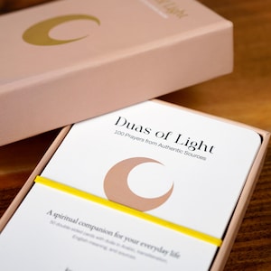 K&ouml;nnte beinhalten: Ein rosafarbenes Box-Set mit dem Schriftzug "Duas of Light" und einem Halbmond-Design. Die Box enth&auml;lt Karten mit dem gleichen Design, die von einem gelben Gummiband zusammengehalten werden. Der Text lautet "100 Gebete aus authentischen Quellen".