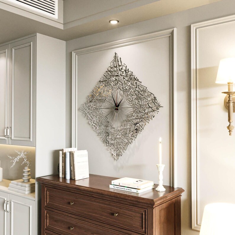 May include: A decorative wall clock with a diamond-shaped design. The clock features intricate, stylized Arabic calligraphy in a silver finish. The clock hands are black. The clock is mounted on a white wall above a wooden dresser.
