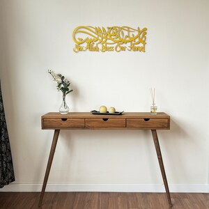 May include: A gold-colored wooden wall decor with Arabic calligraphy that reads "Ya Allah Bless Our Home". The decor is hanging on a white wall above a wooden console table with three drawers. The table has a black tray with two yellow decorative spheres on top.