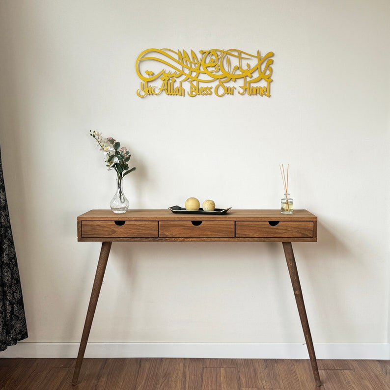 May include: A gold-colored wooden wall decor with Arabic calligraphy that reads "Ya Allah Bless Our Home". The decor is hanging on a white wall above a wooden console table with three drawers. The table has a black tray with two yellow decorative spheres on top.