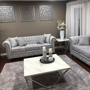 May include: A gray tufted sofa with two throw pillows sits in front of a wall with three metal wall decor pieces. A white marble coffee table with a silver metal frame sits in front of the sofa. The coffee table has a black tray with a silver metal orb and two white candles.