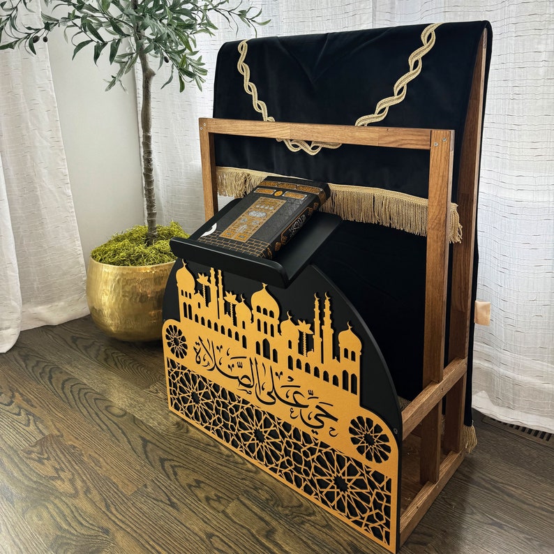 May include: A wooden prayer stand with a black velvet cushion and a gold-colored metal design. The stand features a book with Arabic text and a decorative cutout of a mosque.