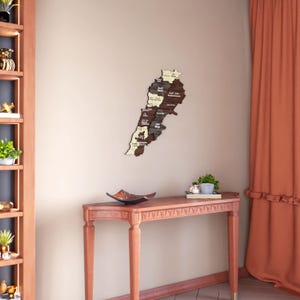 May include: A wooden wall art piece depicting the map of Lebanon with the names of each region written in Arabic. The map is mounted on a beige wall above a red console table with a decorative black tray.