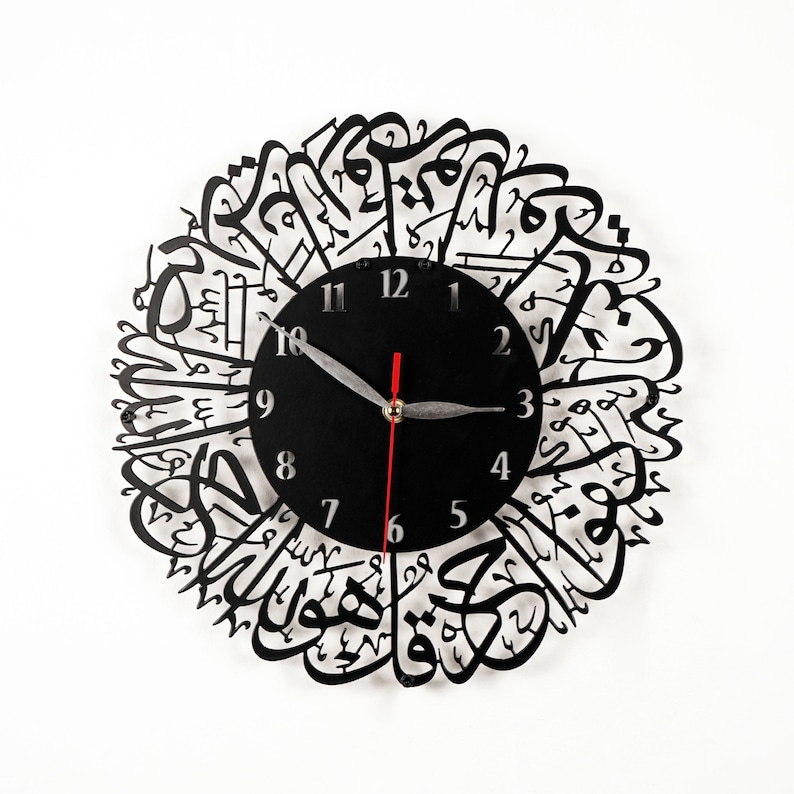 K&ouml;nnte beinhalten: Schwarze Wanduhr mit arabischer Kalligraphie, die das Zifferblatt umgibt. Das Zifferblatt hat schwarze Zeiger und wei&szlig;e Zahlen.