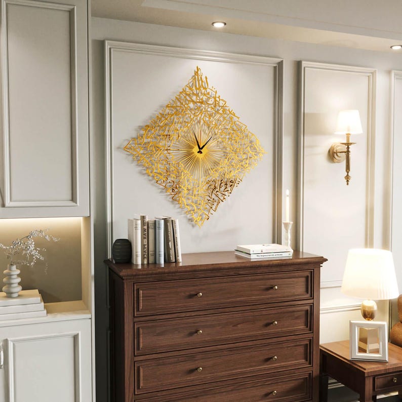 May include: A gold-colored, diamond-shaped wall clock with Arabic calligraphy. The clock has a sunburst design and black clock hands. The clock is mounted on a light gray wall above a dark brown dresser.