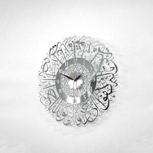 May include: A silver wall clock with Arabic calligraphy around the face. The clock face has black hands and black numbers.