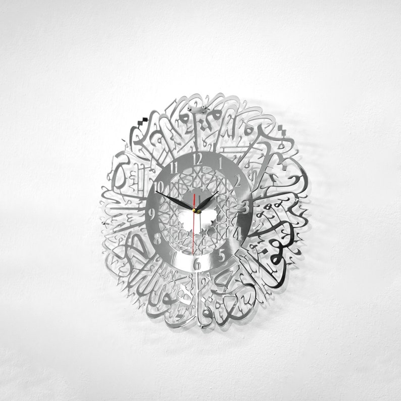 May include: A silver wall clock with Arabic calligraphy around the face. The clock face has black hands and black numbers.