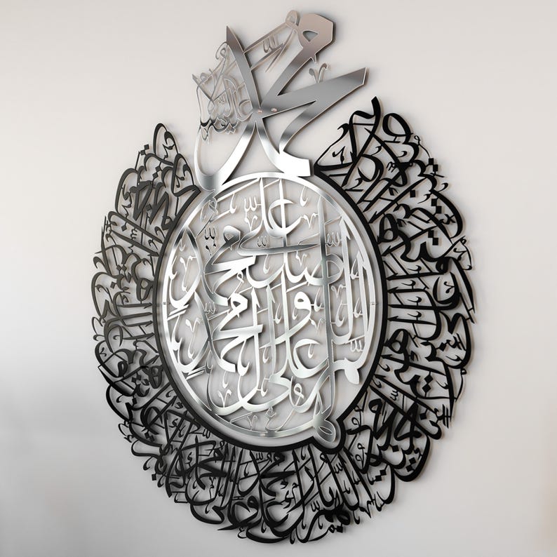 May include: A silver and black metal wall art piece featuring intricate Arabic calligraphy. The design is circular with a central circle containing a larger inscription surrounded by a ring of smaller inscriptions.