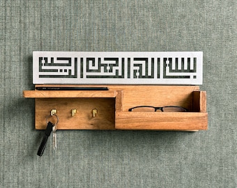 May include: A wooden key holder with a metal plate featuring Arabic calligraphy. The holder has three hooks and a small shelf for glasses or other items.