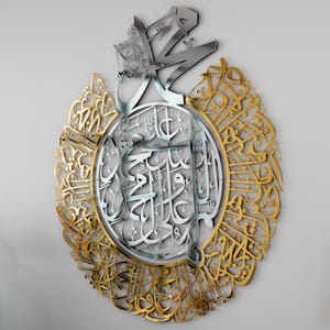 May include: A silver and gold metal wall art featuring intricate Arabic calligraphy. The design is circular with a central silver section and a gold outer ring. The calligraphy is in a traditional style and is likely a religious inscription.