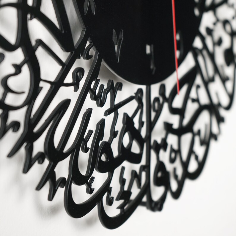 K&ouml;nnte beinhalten: Schwarze Uhr mit arabischer Kalligrafie. Das Zifferblatt ist schwarz mit wei&szlig;en Zahlen und einem roten Sekundenzeiger.