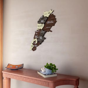 May include: A wooden map of Lebanon with the names of the different regions in Arabic and English. The map is mounted on a beige wall above a wooden table with a decorative bowl and a potted plant.