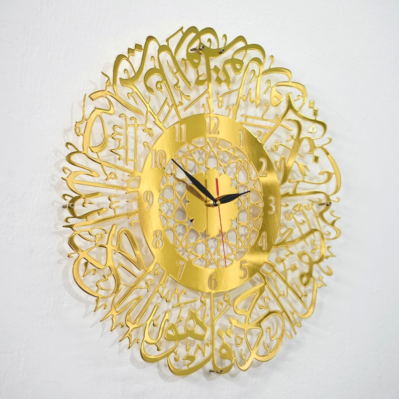 May include: Gold-colored wall clock with Arabic calligraphy surrounding the clock face. The clock face has black hands and Roman numerals.