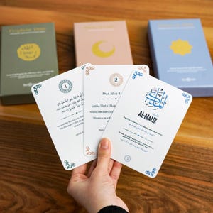Peut inclure: Une main tient trois cartes blanches avec des écritures arabes et des bordures décoratives, avec trois coffrets en arrière-plan. Les cartes portent les textes "AL-MALIK" et "Dua After E". Les boîtes sont vertes, roses et bleues.