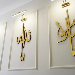 May include: Three framed wall art pieces with gold calligraphy on a white background. Each piece has a gold light fixture above it. The calligraphy is in a flowing script style.