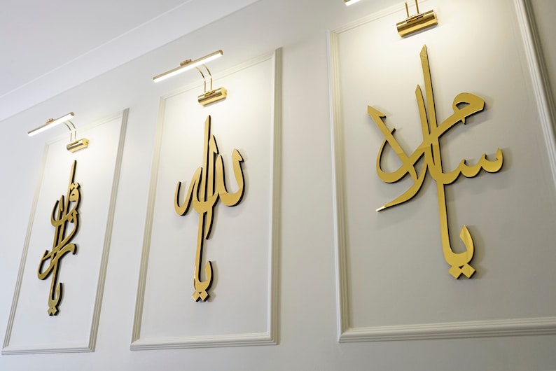 May include: Three framed wall art pieces with gold calligraphy on a white background. Each piece has a gold light fixture above it. The calligraphy is in a flowing script style.