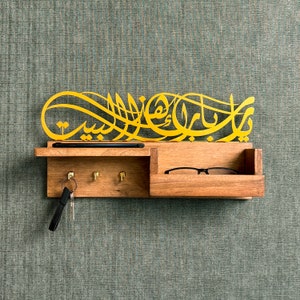 Op de afbeelding: Een houten wandbevestigde sleutelhanger met een gouden metalen ontwerp met Arabische kalligrafie. De houder heeft drie haken voor sleutels en een klein plankje voor brillen of een telefoon.