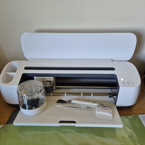May include: A white Cricut cutting machine with the lid open, revealing the interior. A clear jar and several tools sit on the tray. The machine is on a wooden surface with a green cutting mat visible.