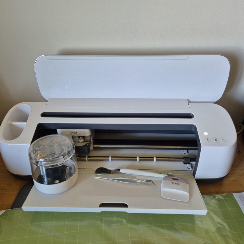 May include: A white Cricut cutting machine with the lid open, revealing the interior. A clear jar and several tools sit on the tray. The machine is on a wooden surface with a green cutting mat visible.