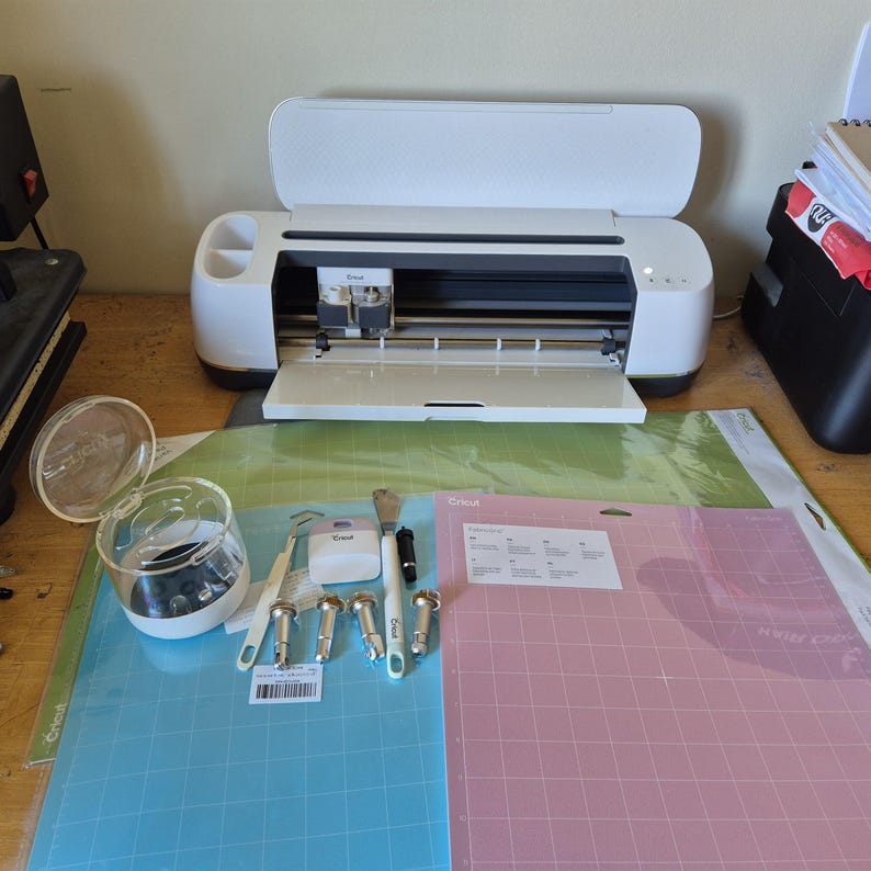 May include: A white Cricut cutting machine with an open lid, surrounded by crafting tools and mats. Includes a blue and pink cutting mat, a clear container, and various tools with white handles.