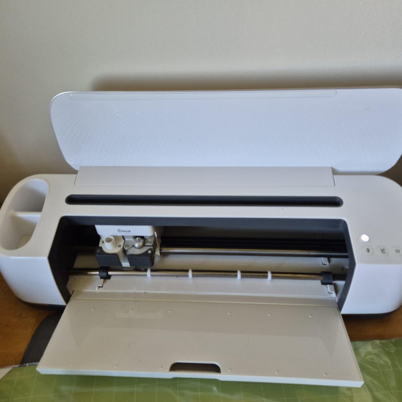 May include: A white Cricut cutting machine with the lid open, revealing the blade and cutting area. The machine has a sleek design with a built-in storage compartment on the left side. The front panel is open, and a green cutting mat is visible.