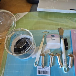 May include: A collection of Cricut crafting tools and accessories, including a clear plastic dome, tweezers, a scraper, and various cutting blades. The items are arranged on a blue and green cutting mat.