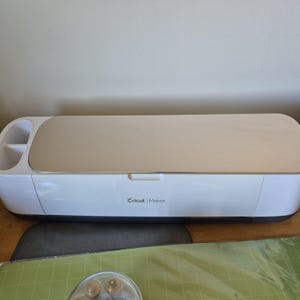May include: A white Cricut Maker machine with a beige top and the Cricut Maker logo. The machine is on a wooden surface with a gray mat and green cutting mat.