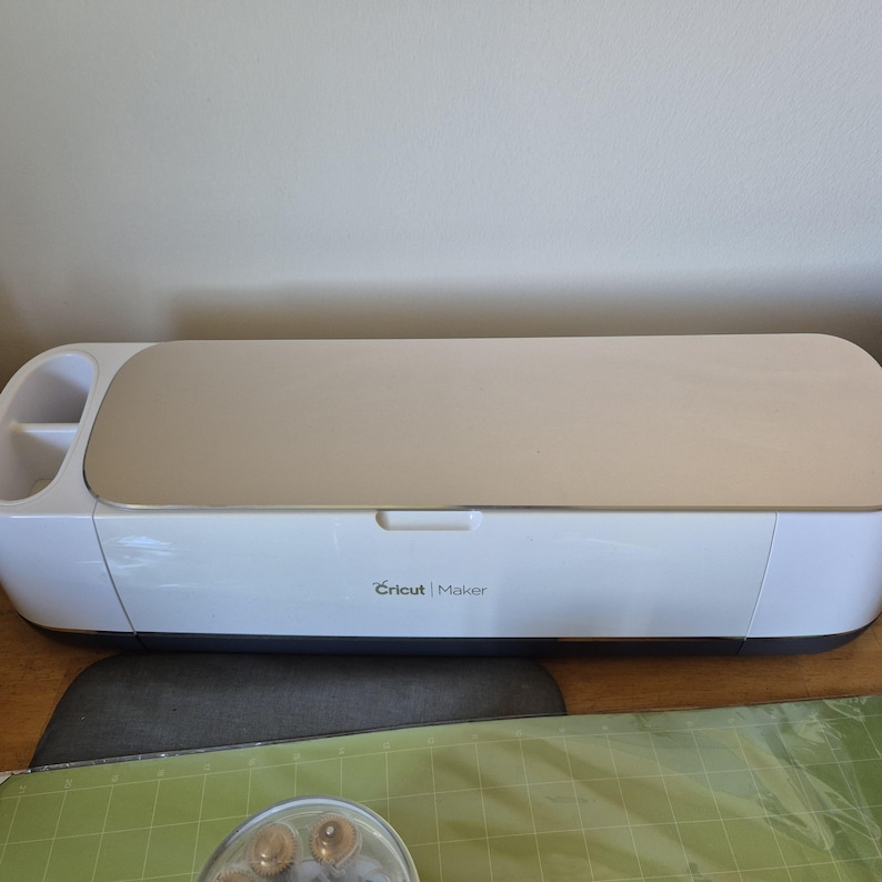 May include: A white Cricut Maker machine with a beige top and the Cricut Maker logo. The machine is on a wooden surface with a gray mat and green cutting mat.
