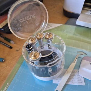 May include: A clear plastic Cricut tool organizer with a lid, containing several gold and silver-colored tools. A silver tweezer and a pink and white scraper are also visible, along with a blue cutting mat.