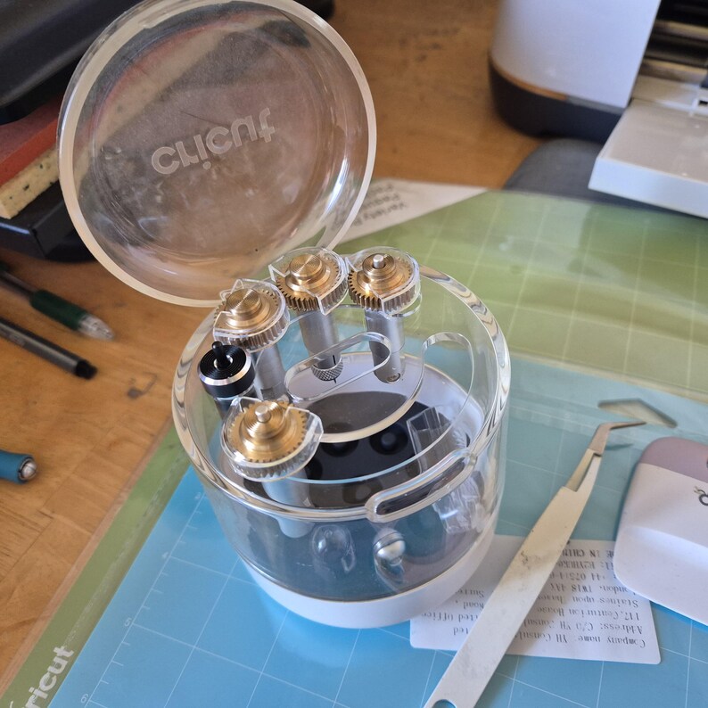 May include: A clear plastic Cricut tool organizer with a lid, containing several gold and silver-colored tools. A silver tweezer and a pink and white scraper are also visible, along with a blue cutting mat.