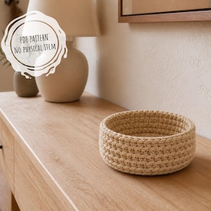 Könnte beinhalten: Ein runder, beigefarbener Häkelkorb mit einem strukturierten Rippenmuster. Das Bild enthält eine weiße, kreisförmige Grafiküberlagerung mit dem schwarzen Text "PDF PATTERN NO PHYSICAL ITEM", was darauf hinweist, dass es sich um eine digitale Datei handelt.