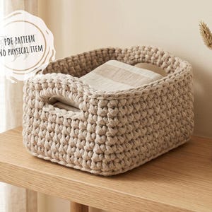 Puede incluir: Una cesta rectangular de ganchillo beige con asas, que contiene tela doblada. La cesta está hecha de hilo grueso y se encuentra sobre una superficie de madera. El texto "PDF PATTERN NO PHYSICAL ITEM" está en la esquina superior izquierda.