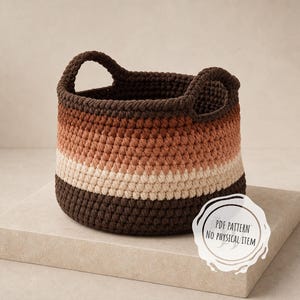 May include: A round crochet basket featuring horizontal stripes in dark brown, terracotta, and cream. The basket has two integrated handles and a textured stitch pattern. A white circular label on the front reads PDF PATTERN NO PHYSICAL ITEM.