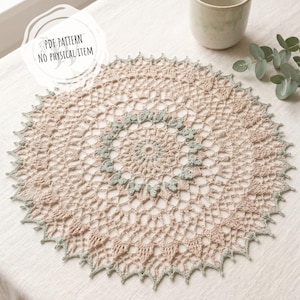 Könnte beinhalten: Handgefertigte, beige-grüne Häkel-Tischdecke mit detailliertem Lochmuster. Die Tischdecke ist kreisförmig, mit einem gewellten Rand und einem zentralen Blumenmuster. Eine Keramiktasse und Grünzeug befinden sich im Hintergrund. Der Text "PDF PATTERN NO PHYSICAL ITEM" befindet sich im Bild.