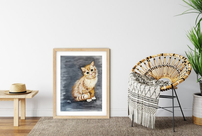 May include: A framed watercolor painting of a ginger kitten sitting on a gray background. The kitten has white markings on its face and chest. The frame is made of wood and has a light brown finish.