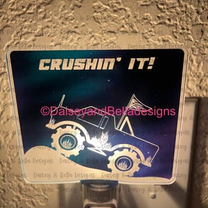 Puede incluir: Una luz nocturna con un fondo degradado azul y morado que presenta una silueta blanca de un camión monstruo con el texto "CRUSHIN' IT!" y el símbolo de copyright seguido de "DaiseyandBeladesigns".