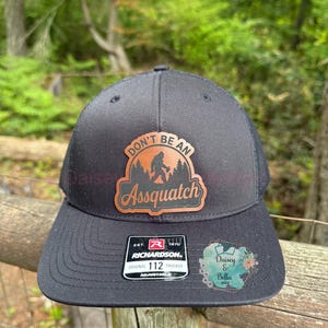 Op de afbeelding: Zwarte truckerpet met een gebogen klep en mesh achterkant. De pet heeft een patch met de tekst "DON'T BE AN Assquatch" en een silhouet van Bigfoot. Het Richardson-merklabel is zichtbaar.