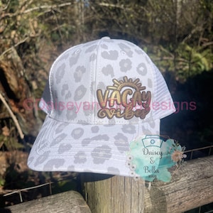 Op de afbeelding: Witte truckercap met een grijs luipaardprint ontwerp. De pet heeft een gouden patch met de tekst "Vacay Vibes" en een zon-graphic. De pet zit op een houten paal.