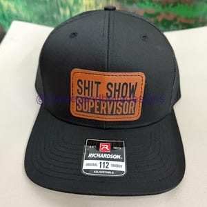 May include: Black Richardson 112 trucker hat with a brown leather patch that reads "SHIT SHOW SUPERVISOR". The hat has a mesh back and an adjustable strap. The Richardson brand tag is visible.