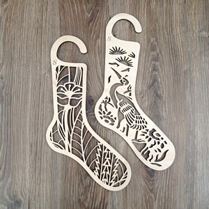 May include: Two wooden stocking-shaped ornaments with intricate cut-out designs. One features a floral pattern, the other a crane with a sunrise. Each has a curved top for hanging and an "S" initial.