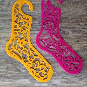 Può includere: Due appendini di plastica a forma di calza con un motivo floreale. L'appendino a sinistra è giallo e l'appendino a destra è rosa. Entrambi gli appendini hanno una piccola 'S' stampata su di essi.